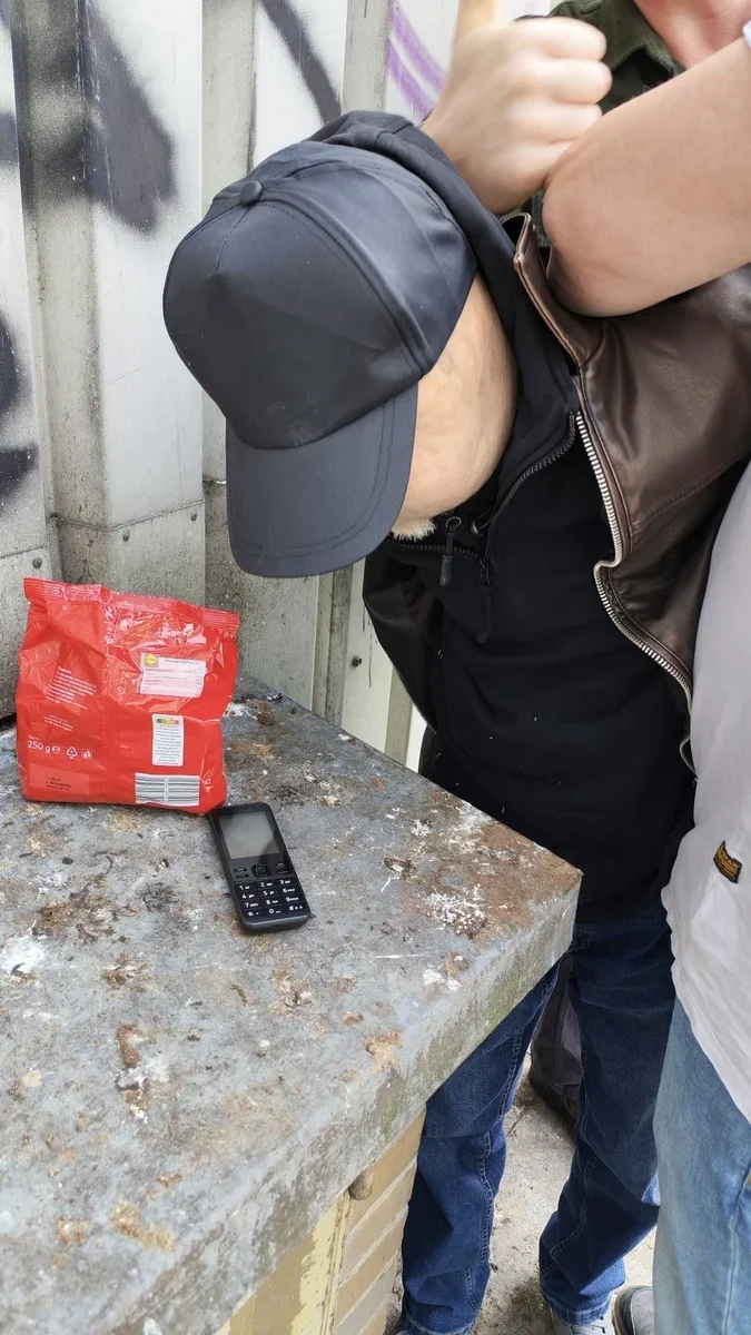 Mężczyzna w czarnej czapce i skórzanej kurtce pochyla się nad betonowym blatem, na którym leży czerwona paczka i telefon komórkowy.