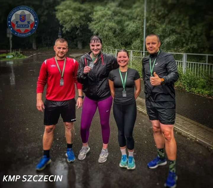 Czterech biegaczy z medalami na szyjach pozuje na mokrej asfaltowej ścieżce w parku, w lewym górnym rogu widoczny herb Komendy Miejskiej Policji w Szczecinie.