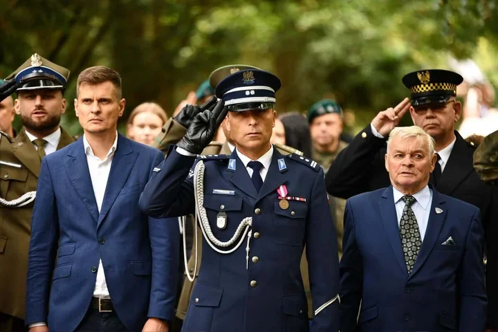 Uroczystość z udziałem funkcjonariuszy mundurowych i mężczyzn w garniturach, podczas której policjant w granatowym mundurze salutuje.