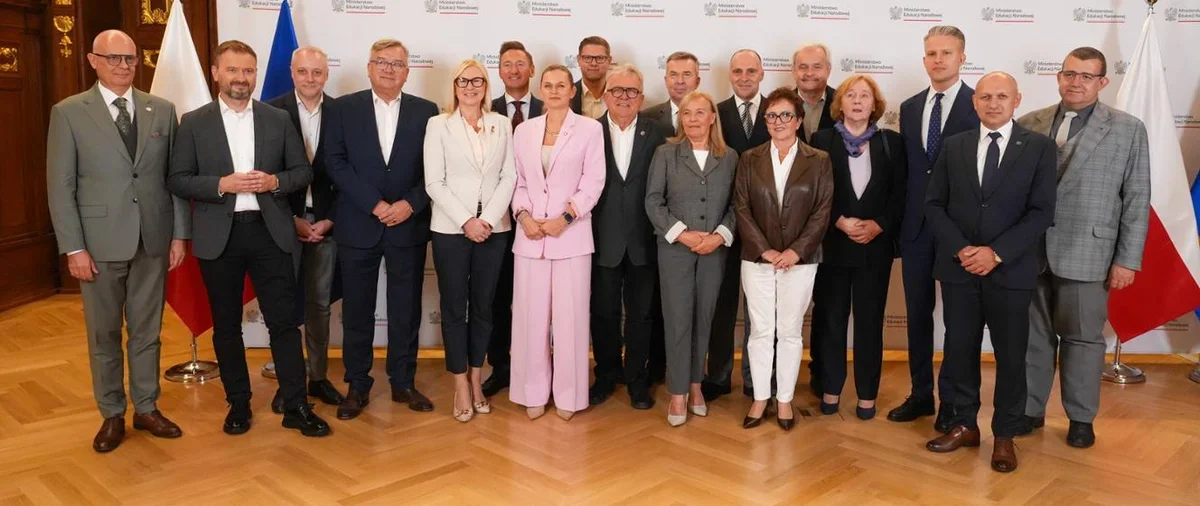 Grupa osób w eleganckim stroju biznesowym pozuje do pamiątkowego zdjęcia w pomieszczeniu reprezentacyjnym przed ścianką z logotypami Ministerstwa Edukacji Narod
