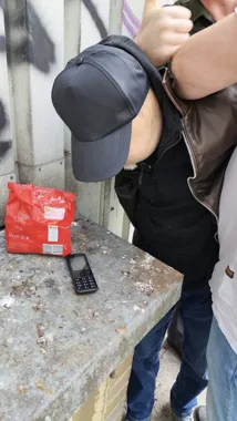 Mężczyzna w czarnej czapce i skórzanej kurtce pochyla się nad betonowym blatem, na którym leży czerwona paczka i telefon komórkowy.