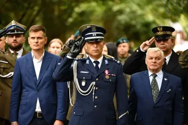 Uroczystość z udziałem funkcjonariuszy mundurowych i mężczyzn w garniturach, podczas której policjant w granatowym mundurze salutuje.