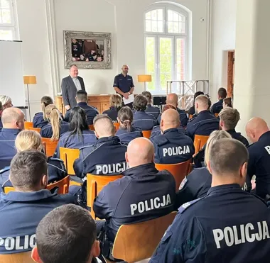Policjanci w mundurach siedzą na krzesłach w sali i słuchają dwóch mężczyzn stojących z przodu, jednego w garniturze, drugiego w policyjnym mundurze.