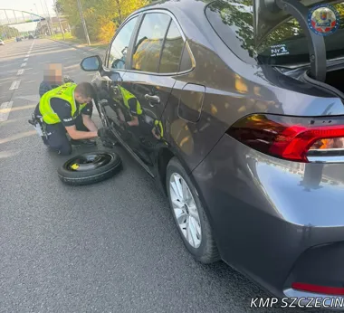 Policjant w kamizelce odblaskowej wymienia koło w szarej Toyocie Corolla na jezdni w Szczecinie.