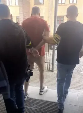 Mężczyzna w czerwonej koszulce i szortach prowadzony jest w kajdankach przez dwóch funkcjonariuszy w kierunku wyjścia z budynku.
