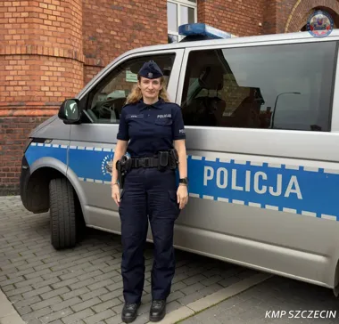 Policjantka w mundurze stoi przed policyjnym radiowozem na tle ceglanego budynku.