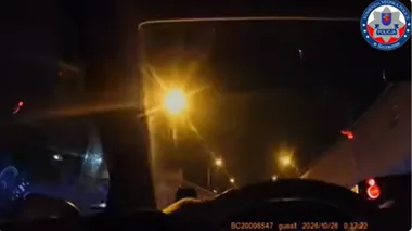 Nocny kadr z policyjnej kamery samochodowej pokazujący ulicę oświetloną latarniami i pojazdy w ruchu.