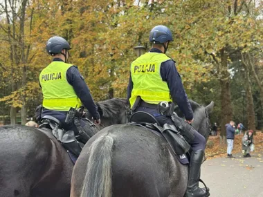 Dwóch policjantów na koniach patroluje parkową alejkę w jesienny dzień.