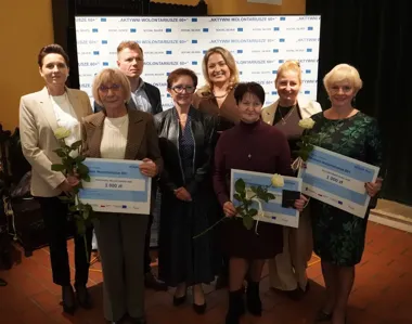 Grupa osób pozuje do zdjęcia podczas uroczystości wręczenia nagród w programie „Aktywni Wolontariusze 60+”, trzy laureatki trzymają czeki na 1000 zł i białe róż