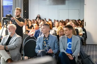 Publiczność siedzi na sali konferencyjnej podczas wydarzenia, a w tle kamerzysta nagrywa spotkanie.