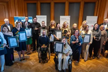 Grupa osób z dyplomami i nagrodami pozuje do pamiątkowego zdjęcia podczas uroczystej gali w sali z drewnianą boazerią.