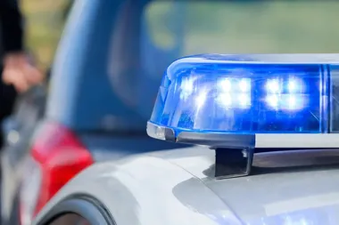 Zbliżenie na niebieski sygnał świetlny na dachu policyjnego radiowozu, w tle rozmyta sylwetka osoby i inny samochód.