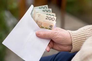 Starsza osoba w beżowym swetrze trzyma białą kopertę z banknotami 100 i 200 złotych na rozmytym tle zewnętrznym.