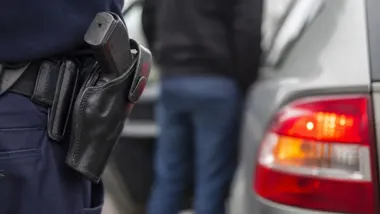 Fragment munduru policjanta z kaburą przy tylnym zderzaku samochodu osobowego.