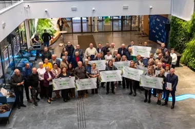 Grupa osób pozuje do pamiątkowego zdjęcia w nowoczesnym holu, trzymając duże czeki z napisami „Przyjazne ogrody 2026” i kwotami w złotych.