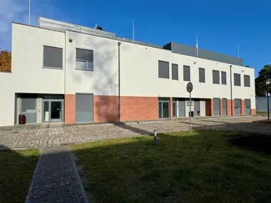 Nowoczesny dwupiętrowy budynek z ceglanym pasem na parterze, otoczony brukowanym placem i trawnikiem, w słoneczny dzień.