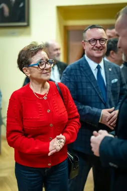 Kobieta w czerwonym swetrze i mężczyzna w granatowej kraciastej marynarce stoją w pomieszczeniu podczas spotkania towarzyskiego.