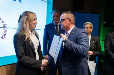 Kobieta w czarnym garniturze i mężczyzna w niebieskim marynarce witają się uściskiem dłoni podczas uroczystości wręczenia dokumentu w sali konferencyjnej.