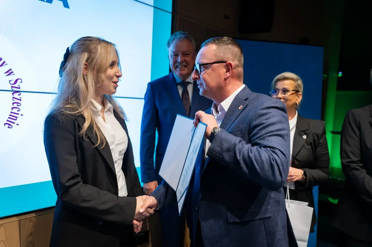 Kobieta w czarnym garniturze i mężczyzna w niebieskim marynarce witają się uściskiem dłoni podczas uroczystości wręczenia dokumentu w sali konferencyjnej.